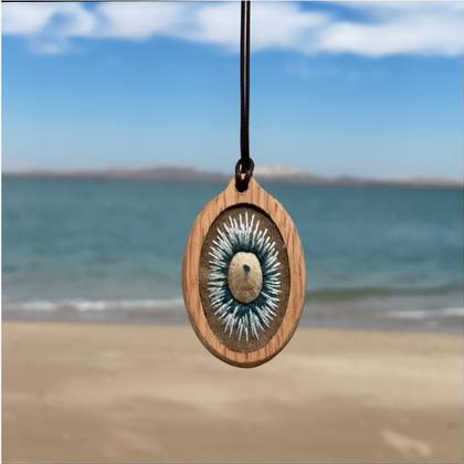 Oval pendant with blue-and-white radial stitching around a pale central oval, photographed at the beach.