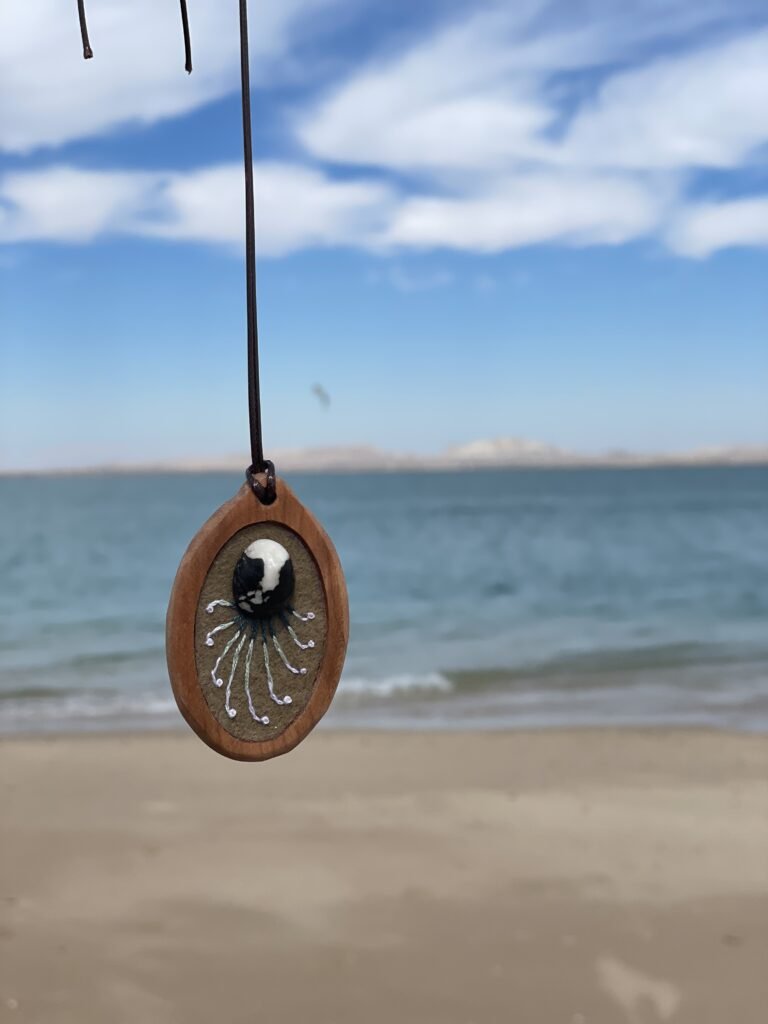 Oval pendant with a black-and-white central focal form and fine pale stitches radiating outward, photographed at the beach.