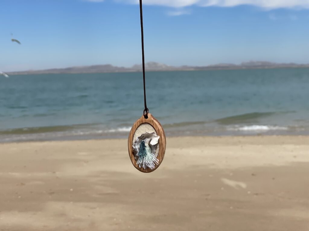 Oval pendant with a pale shell-like focal form above a blue-and-white fan of stitches, hanging against a beach background.