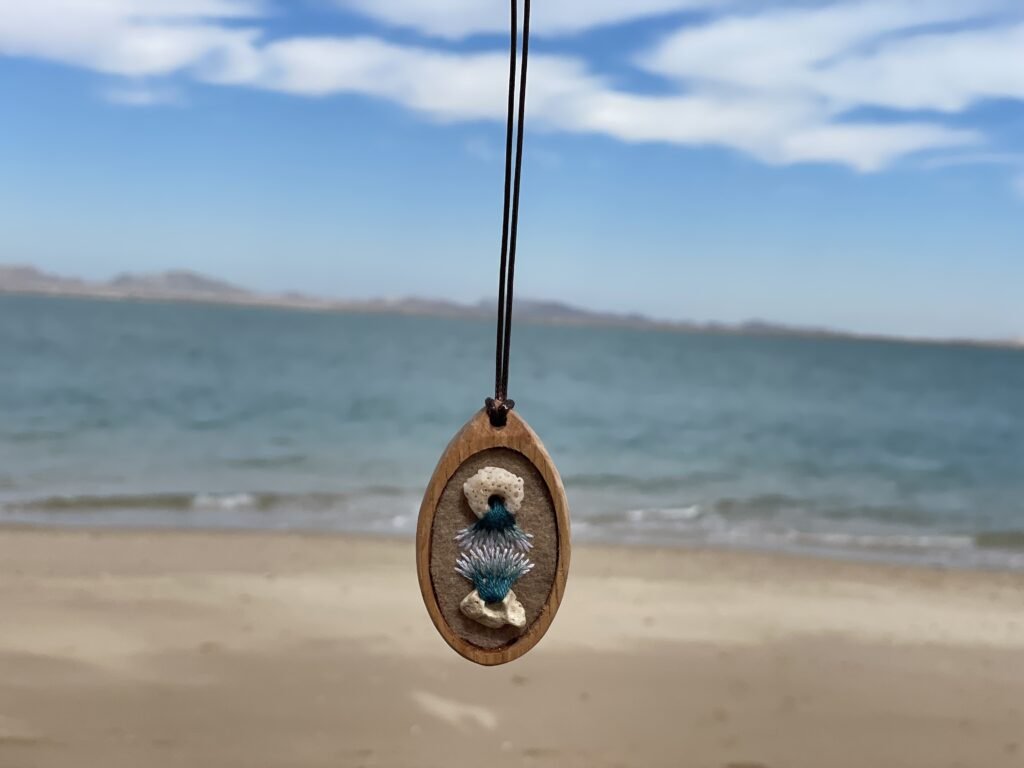 Oval pendant with two pale rounded forms and a teal-and-white current-like embroidered shape between them, photographed at the beach.