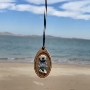 Oval pendant with two pale rounded forms and a teal-and-white current-like embroidered shape between them, photographed at the beach.