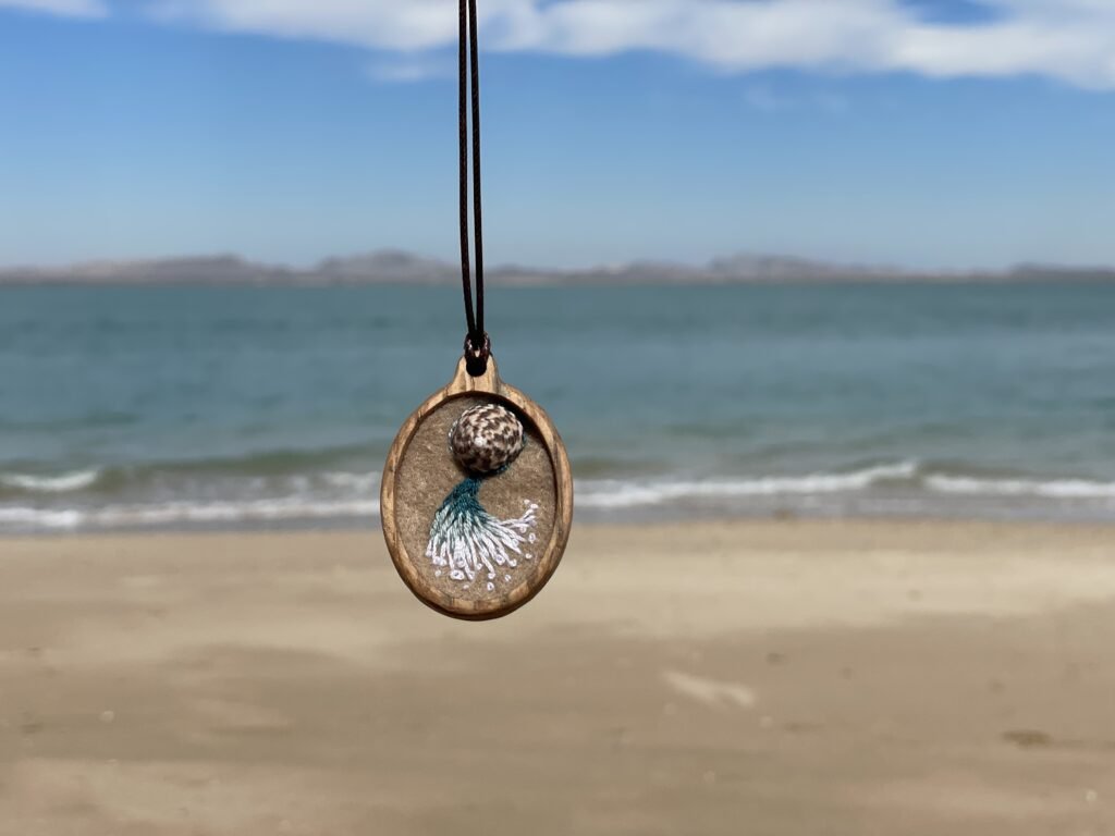 Oval pendant with a mottled round focal detail above a pale teal-and-white fan of stitches, photographed against a beach background.