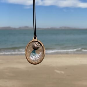 Oval pendant with a mottled round focal detail above a pale teal-and-white fan of stitches, photographed against a beach background.