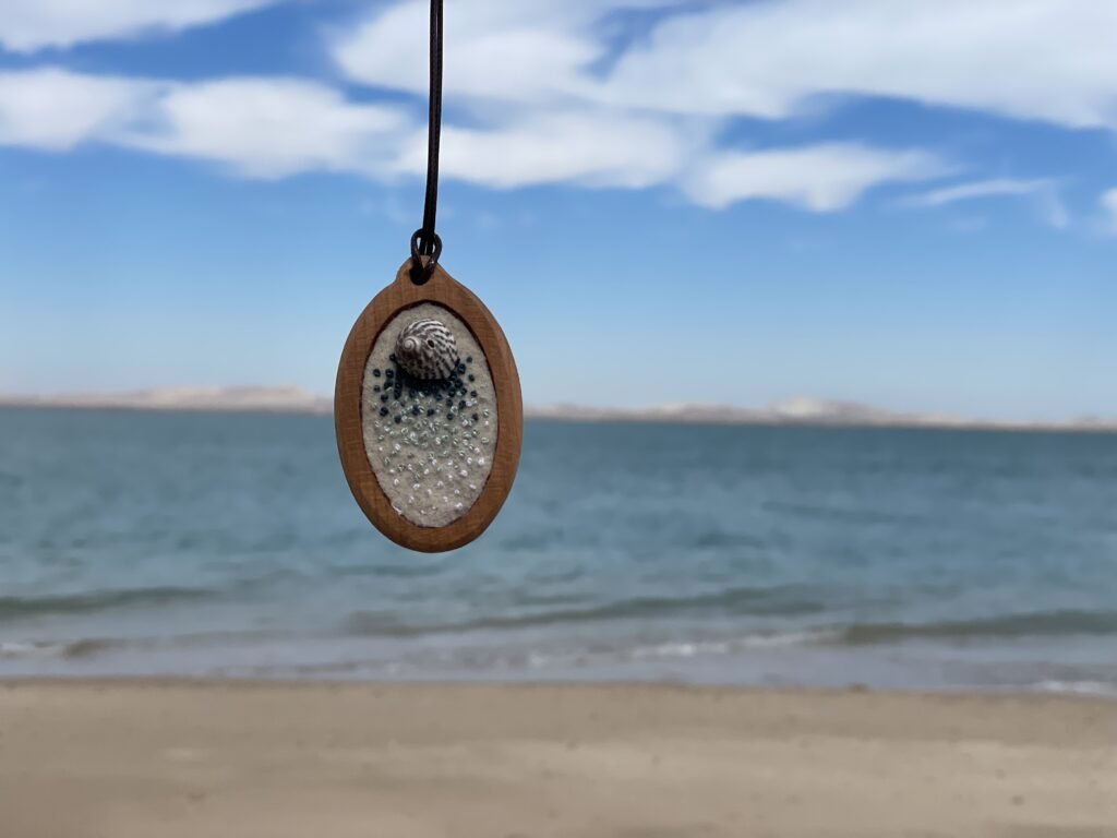 Oval pendant with a mottled focal form and small dark-blue and pale dot-like stitch details, hanging against the beach.
