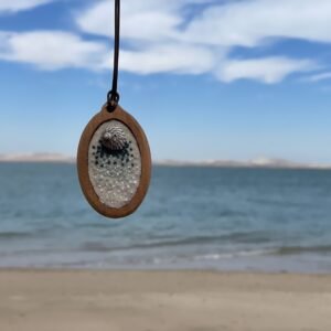 Oval pendant with a mottled focal form and small dark-blue and pale dot-like stitch details, hanging against the beach.