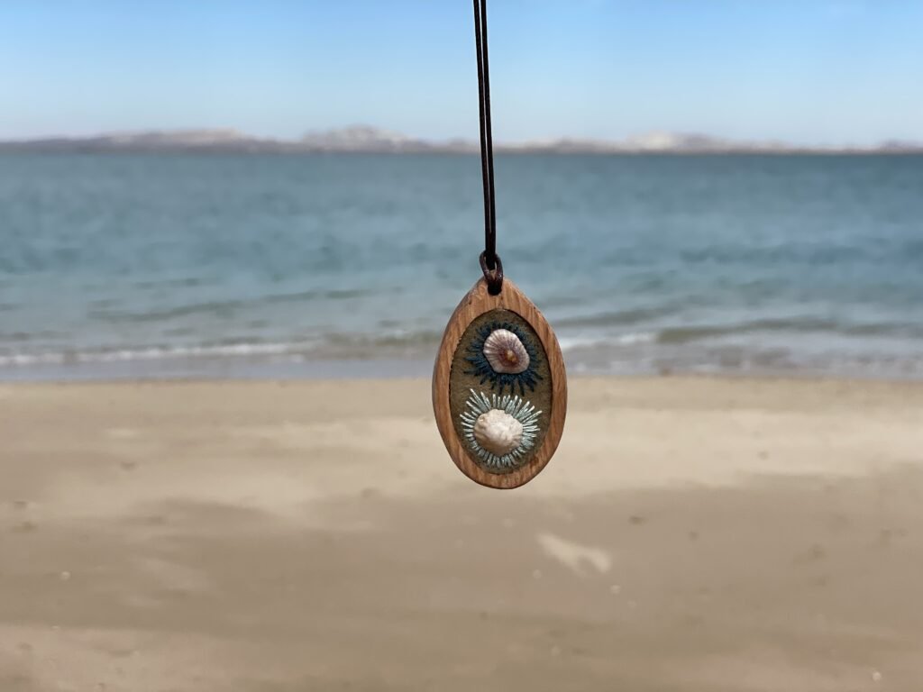 Oval pendant with two pale raised forms framed by dark-blue-and-white radial stitching, hanging against the beach.