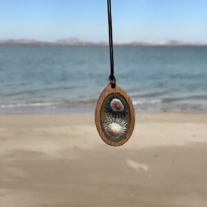 Oval pendant with two pale raised forms framed by dark-blue-and-white radial stitching, hanging against the beach.